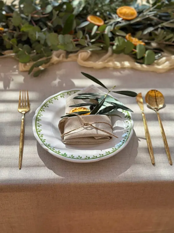 A place setting with a green-rimmed plate, olive sprig, dried orange and gold cutlery on linen