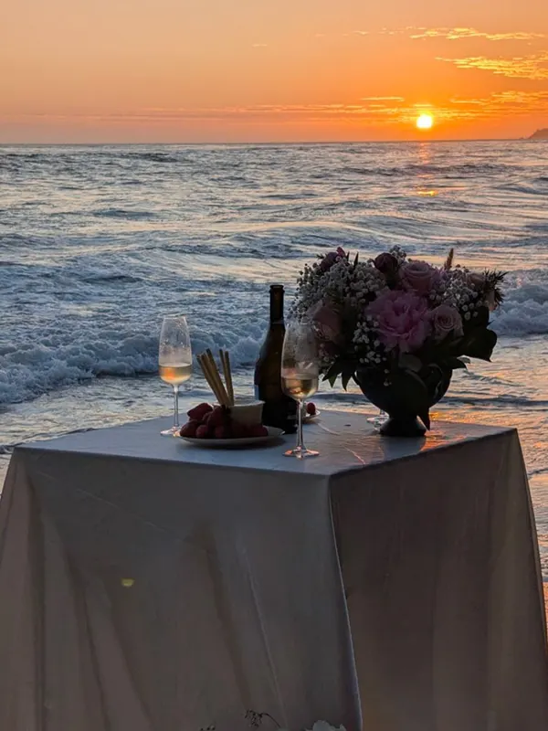 A seaside dinner table with champagne and a bouquet as the sun sets over the ocean