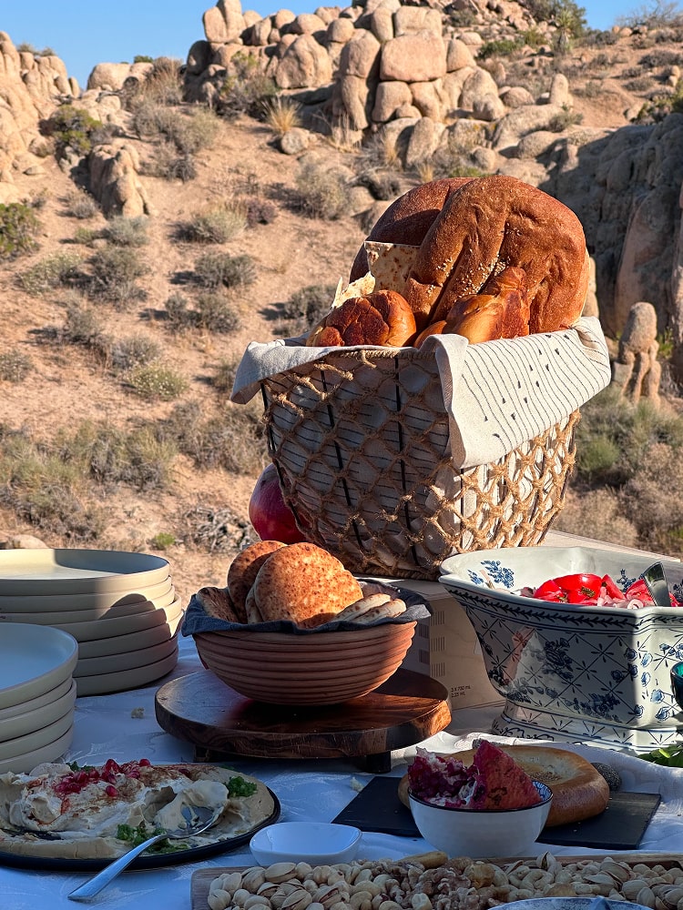 An outdoor feast among desert boulders, with bread in a woven basket, mezze and blue-and-white ceramics