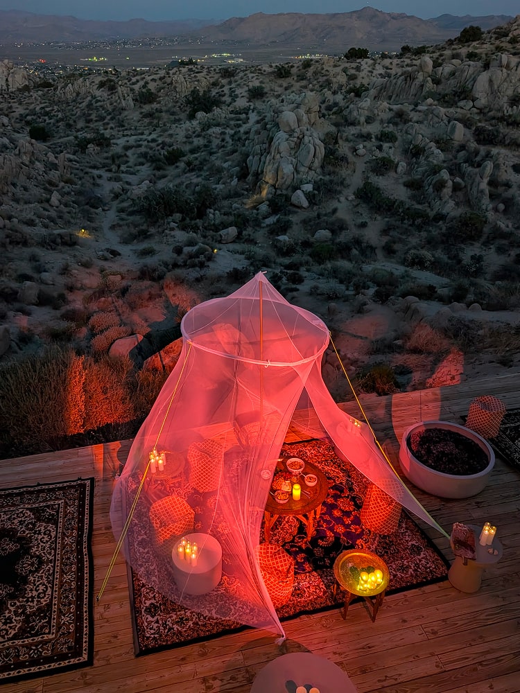 A glowing canopy with lanterns and rugs on a wooden deck overlooking a desert valley at dusk