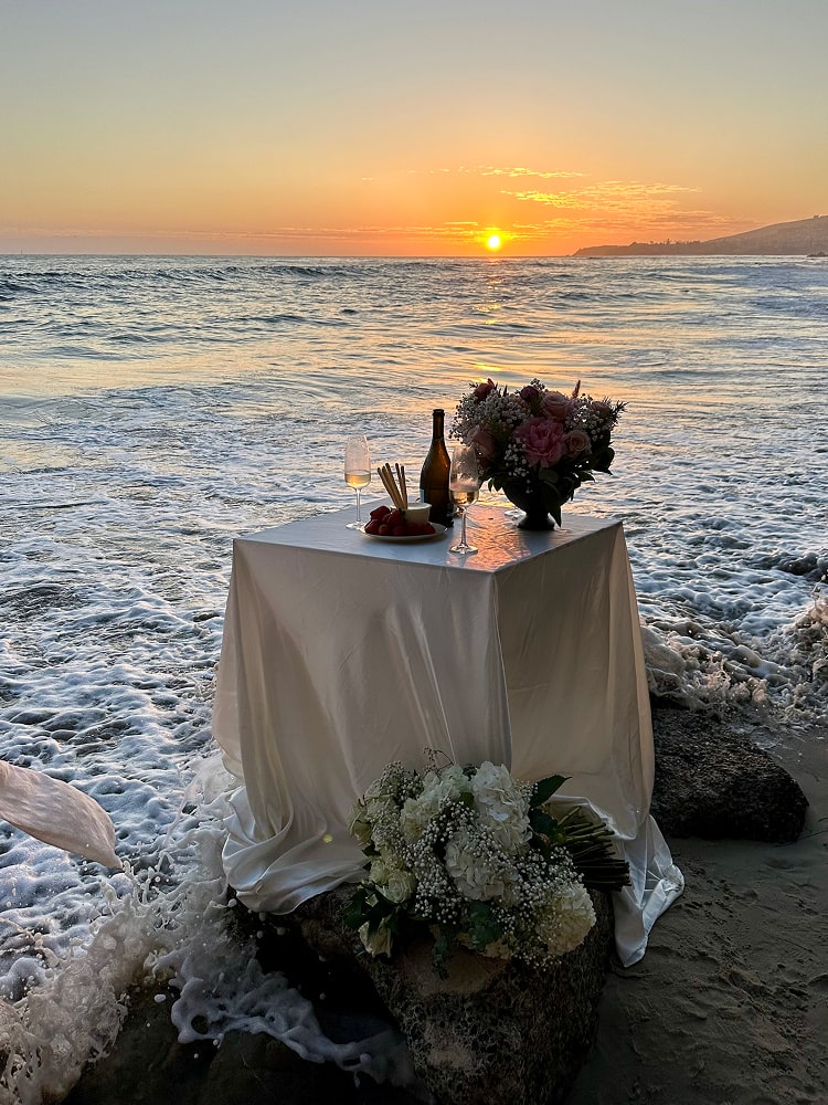 A small draped table with champagne and a bouquet on the rocks at the ocean's edge at sunset