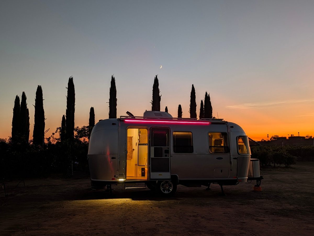 The Silver Hour Airstream at blue hour among cypress trees, door open and lit from within, pink light along the awning